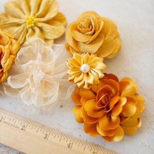 K&ouml;nnte beinhalten: Sortiment gelber Stoffblumen, darunter eine Rose, eine G&auml;nsebl&uuml;mchen und eine Nelke. Die Blumen sind auf einer wei&szlig;en Oberfl&auml;che mit einem Holzlineal angeordnet.