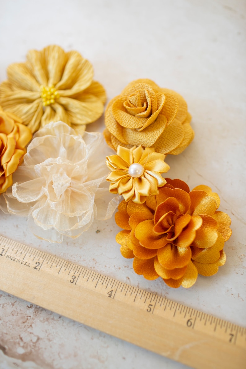 K&ouml;nnte beinhalten: Sortiment gelber Stoffblumen, darunter eine Rose, eine G&auml;nsebl&uuml;mchen und eine Nelke. Die Blumen sind auf einer wei&szlig;en Oberfl&auml;che mit einem Holzlineal angeordnet.