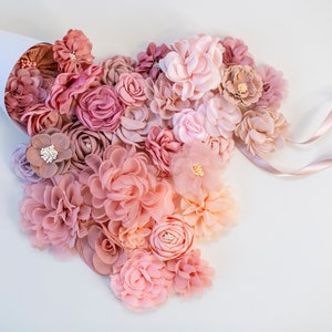 Puede incluir: Una colecci&oacute;n de flores de tela rosa y melocot&oacute;n en varios tama&ntilde;os y estilos. Las flores est&aacute;n dispuestas en forma de coraz&oacute;n y se derraman de un cono de papel blanco.