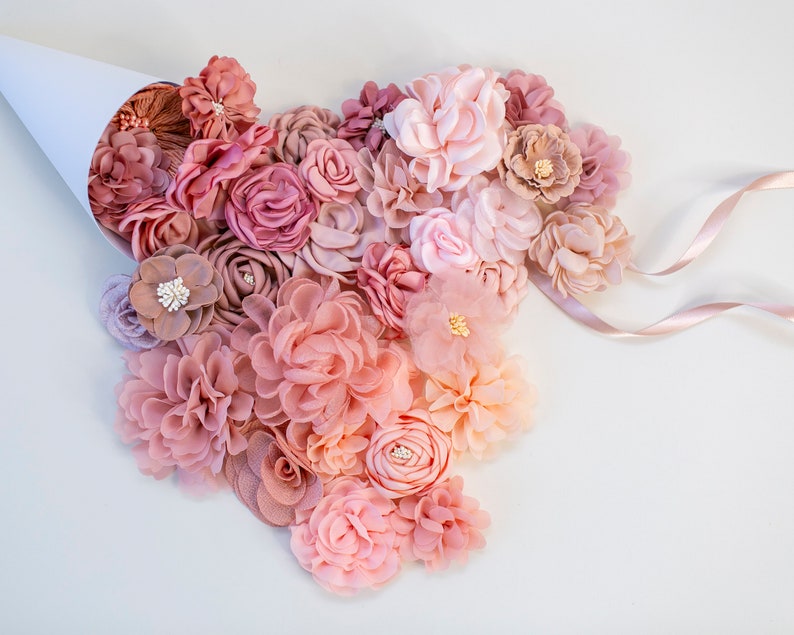 Puede incluir: Una colecci&oacute;n de flores de tela rosa y melocot&oacute;n en varios tama&ntilde;os y estilos. Las flores est&aacute;n dispuestas en forma de coraz&oacute;n y se derraman de un cono de papel blanco.