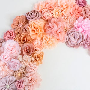 Puede incluir: Una colecci&oacute;n de flores de tela rosa, melocot&oacute;n y marr&oacute;n dispuestas en semic&iacute;rculo sobre un fondo blanco. Las flores est&aacute;n hechas de diferentes materiales y tienen una variedad de texturas.