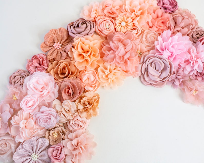 Puede incluir: Una colecci&oacute;n de flores de tela rosa, melocot&oacute;n y marr&oacute;n dispuestas en semic&iacute;rculo sobre un fondo blanco. Las flores est&aacute;n hechas de diferentes materiales y tienen una variedad de texturas.