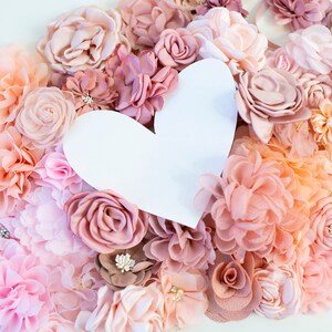 Puede incluir: Un recorte de papel blanco en forma de coraz&oacute;n rodeado de una variedad de flores de tela de color rosa, melocot&oacute;n y crema.