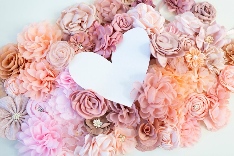 Puede incluir: Un recorte de papel blanco en forma de coraz&oacute;n rodeado de una variedad de flores de tela de color rosa, melocot&oacute;n y crema.