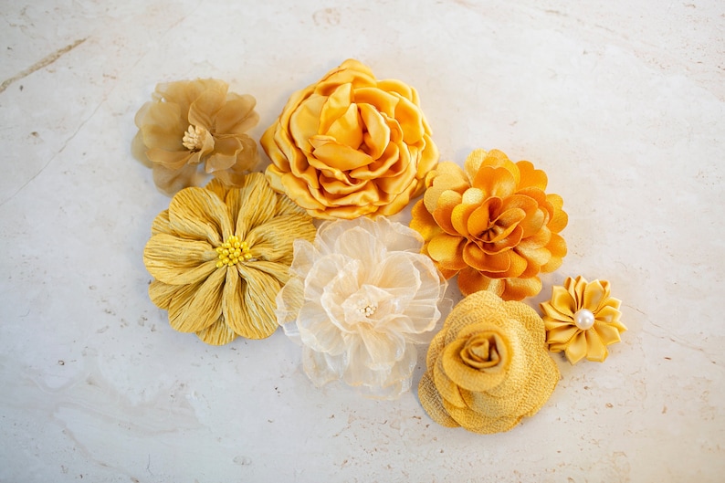 K&ouml;nnte beinhalten: Eine Sammlung von sieben gelben Stoffblumen. Die Blumen sind aus verschiedenen Materialien gefertigt, darunter Satin, Chiffon und T&uuml;ll. Sie sind in einer Gruppe auf einem wei&szlig;en Hintergrund angeordnet.
