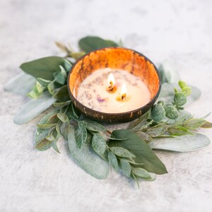 Puede incluir: Una vela blanca con tres mechas ardiendo en un cuenco de c&aacute;scara de coco marr&oacute;n. El cuenco est&aacute; rodeado por una corona de hojas verdes.