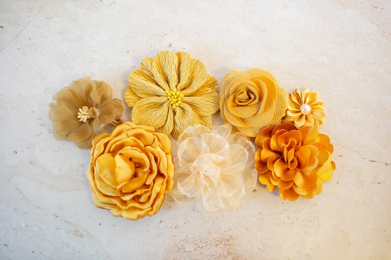K&ouml;nnte beinhalten: Sortiment aus sieben gelben Stoffblumen in verschiedenen Stilen und Texturen. Die Blumen sind auf einem hellbeigen Hintergrund angeordnet.