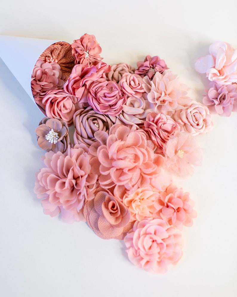 Puede incluir: Una colecci&oacute;n de flores de tela rosa y melocot&oacute;n en varias formas y tama&ntilde;os, que se derraman de un cono de papel blanco.