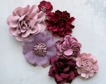 May include: Assortment of seven fabric flowers in shades of pink and burgundy. The flowers are arranged in a cluster on a white background.