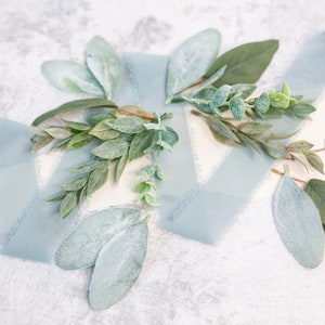 Puede incluir: Un plano de cinta azul y hojas verdes de eucalipto sobre un fondo blanco. Las hojas tienen una ligera capa de polvo blanco.