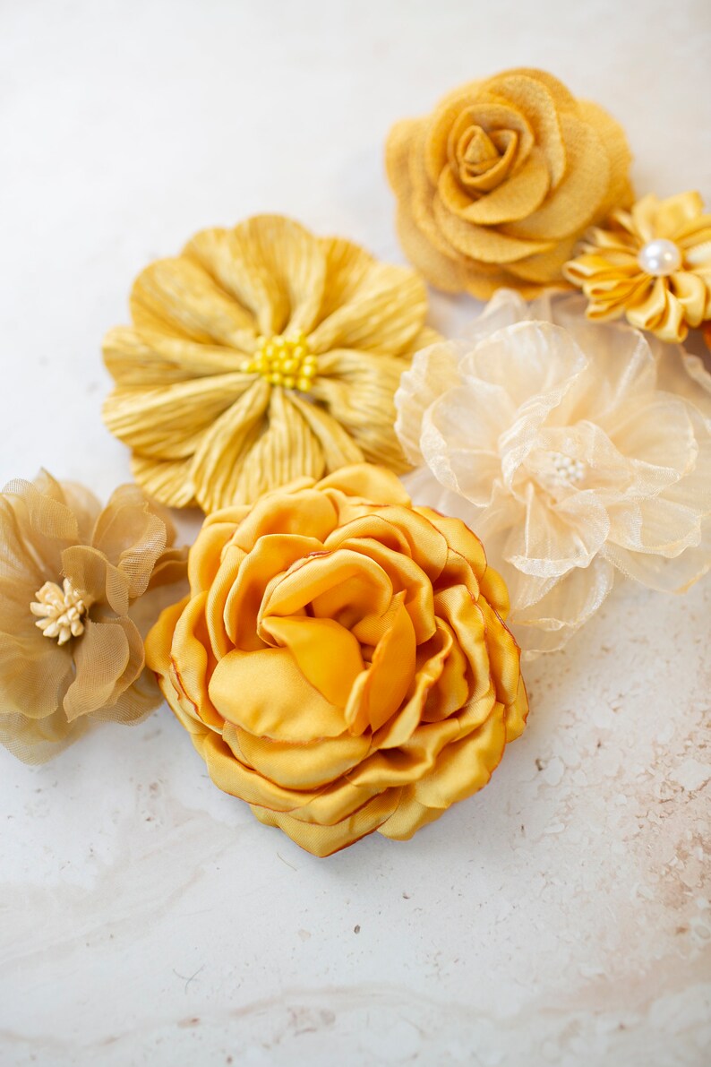 K&ouml;nnte beinhalten: Sortiment gelber Stoffblumen, darunter eine Rose, eine G&auml;nsebl&uuml;mchen und eine gekr&auml;uselte Blume. Die Blumen sind auf einem wei&szlig;en Hintergrund angeordnet.