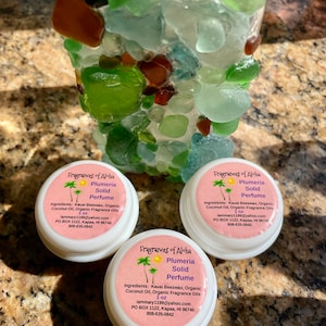 May include: Three white round containers with lids, each containing a solid perfume. The containers are labeled "Fragrances of Aloha Plumeria Solid Perfume" and list the ingredients, contact information, and website. The containers are sitting on a surface with a background of colorful sea glass.