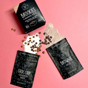 May include: A black baking kit box and pouches are displayed on a pink background. The box reads "BROOKIES BAKING KIT". The pouches say "CHOC CHIP COOKIE MIX" and "BROWNIE MIX". Chocolate chips and flour are also scattered around.