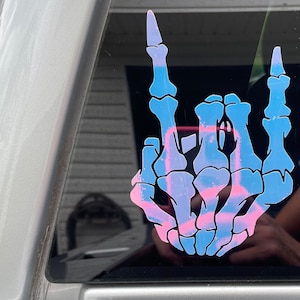 Könnte beinhalten: Eine blaue und rosafarbene Skeletthand, die die Rock'n'Roll-Handgeste macht. Die Hand ist ein Aufkleber auf einer Autoscheibe.