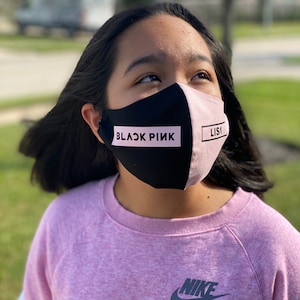 May include: A young person wearing a pink sweatshirt with a black Nike logo and a black and pink face mask with the text "BLACKPINK" and "LISA" on it.
