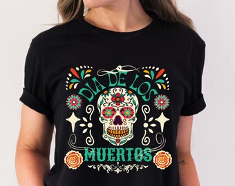 May include: Black t-shirt featuring a colorful sugar skull design with the words "DIA DE LOS MUERTOS" in teal and white. The skull has floral accents in red, orange, and teal. The shirt is a crew neck style.