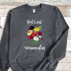 May include: A dark gray sweatshirt with the text "that's just ranunculus" in white lettering. The sweatshirt features a colorful bouquet of ranunculus flowers in the center.