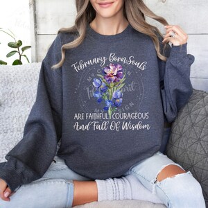 May include: A dark gray sweatshirt with a white graphic design. The graphic features a purple and blue flower with a white circle around it. The text "February Born Souls" is above the flower and the text "My Creation Sign Are Faithful Courageous And Full Of Wisdom" is below the flower.
