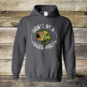 May include: Dark gray hooded sweatshirt with the text "DON'T BE A BASS HOLE" surrounding a cartoon bass fish with its mouth open. The fish is green with red eyes. The sweatshirt has a drawstring hood.