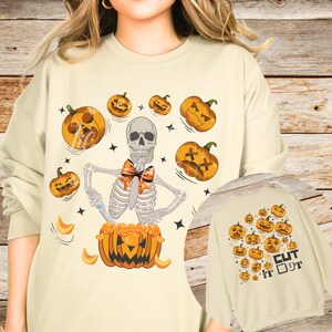 May include: Cream-colored sweatshirt featuring a Halloween-themed design. The graphic includes a skeleton with a bow tie emerging from a carved pumpkin, surrounded by jack-o'-lanterns. The words "Cut It Out" are printed on the back.