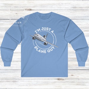 May include: Light blue long-sleeve shirt with the text "I'M JUST A PLANE GUY" and an illustration of a white and gray airplane. The graphic is inside a white circle. The shirt is made of a soft, comfortable material.