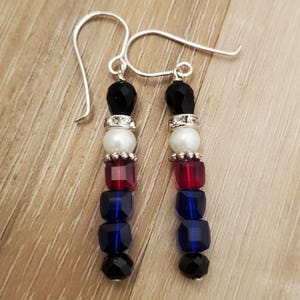 May include: A pair of dangle earrings with black, white, red, and blue beads. The earrings are made with silver hooks.