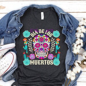 May include: A dark grey t-shirt with a colorful sugar skull design and the text "Dia de los Muertos" in purple lettering.
