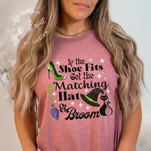 May include: Mauve t-shirt with the text "If the Shoe Fits Get the Matching Hat & Broom." The design includes a green high heel, a witch's hat, a black cat, and a broom. The shirt is decorated with white stars.