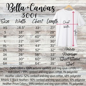 May include: A white Unisex Premium Short Sleeve Shirt from Bella+Canvas, size chart with measurements in inches. The shirt is hanging on a wooden hanger. The chart includes size, width, chest, and length measurements.