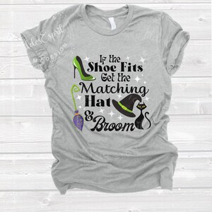 May include: A heather gray t-shirt with a Halloween-themed graphic. The design includes a green high heel, a witch's hat, a black cat, and a broom. The text reads "If the Shoe Fits Get the Matching Hat & Broom."