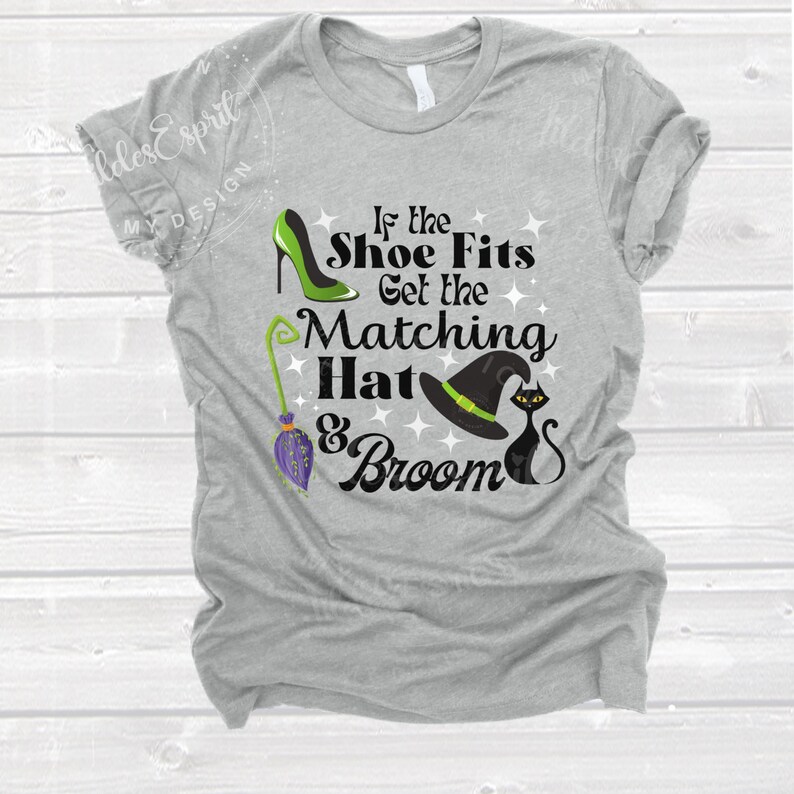 May include: A heather gray t-shirt with a Halloween-themed graphic. The design includes a green high heel, a witch's hat, a black cat, and a broom. The text reads "If the Shoe Fits Get the Matching Hat & Broom."