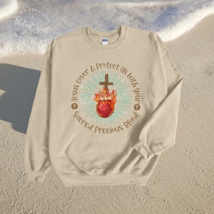 May include: A beige sweatshirt with a graphic design of a flaming heart with a cross on top. The text around the heart reads "Jesus cover & protect us with your sacred precious blood".