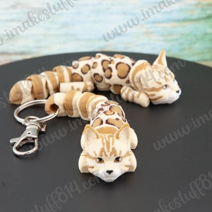 May include: A brown and white spotted cat keychain with a silver key ring. The cat is curled up in a sleeping position. The keychain is made of a soft, flexible material.