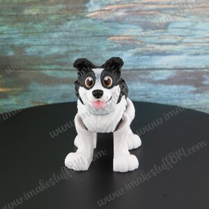 May include: A black and white border collie dog figurine with brown eyes and a pink tongue sticking out. The figurine is standing on a black surface.