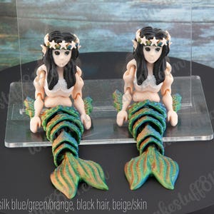 May include: Two mermaid figurines with black hair, wearing white star-shaped crowns and green and blue tails. The mermaids are posed with their hands raised.