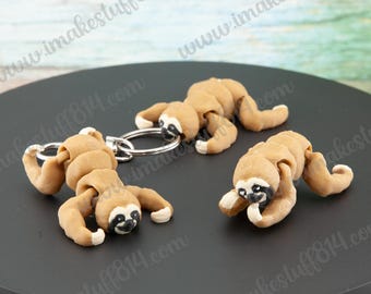 May include: Three brown sloth keychains with black eyes and white accents. The sloths are in different poses, one is curled up, one is hanging upside down, and one is standing on all fours. The keychains are made of polymer clay and are attached to silver key rings.