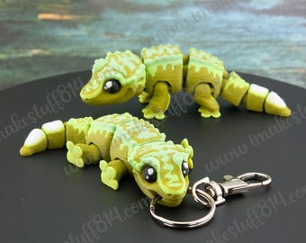 May include: Two articulated lizard figurines featuring a light green and olive green color scheme with white accents on the tail tips. One figurine is attached to a silver metal keychain ring and lobster clasp, while the other is positioned behind it.