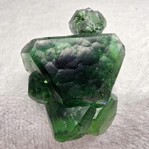 May include: A faceted, green fluorite crystal sculpture. The sculpture has a geometric shape with a large, textured body and a smaller, faceted top. The crystal is a deep, rich green color with some areas of darker green and blue.