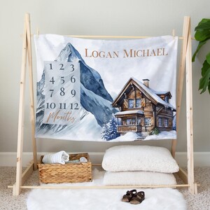 May include: A white baby milestone blanket with a mountain and cabin scene. The blanket has a number chart for each month of the year and the name "Logan Michael" printed on it.