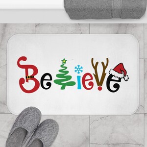 May include: White bath mat with the word "Believe" in colorful letters, decorated with a Christmas tree, snowflake, reindeer antlers, and Santa hat.