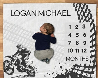 May include: A white baby blanket with a black and white motocross design. The blanket has a black tire track pattern and a black and white illustration of a motocross rider. The blanket also has a black and white number chart for tracking a baby's growth. The text "LOGAN MICHAEL" is printed in black at the top of the blanket.