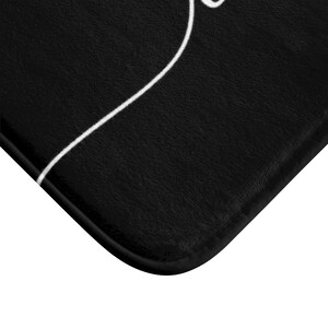 May include: Black bath mat with a white abstract design. The mat has a textured surface and a stitched edge.