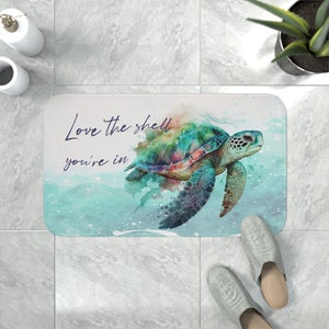 May include: A white bath mat with a watercolor design of a sea turtle swimming in the ocean. The text "Love the shell you're in" is written in purple on the mat.