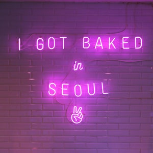 May include: A neon sign on a white tiled wall reads "I got baked in Seoul" with a peace sign below. Two dark brown square stools are in the foreground.