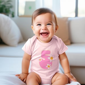 Op de afbeelding: Een lichtroze babyromper met korte mouwen en een grote roze cijfer vijf op de voorkant. Het cijfer is versierd met drie witte madeliefjes met een geel hartje. Het kledingstuk heeft een ronde halslijn en korte mouwen.