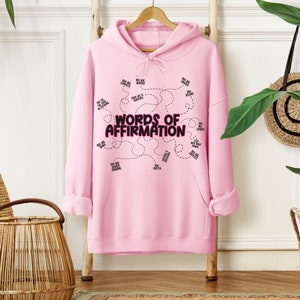 May include: A light pink hooded sweatshirt featuring long sleeves, a front kangaroo pocket, and a drawstring hood. The center graphic displays the text "WORDS OF AFFIRMATION" in bold black letters, surrounded by smaller phrases connected by dashed lines.