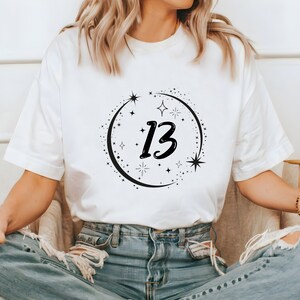 Puede incluir: Una camiseta blanca de manga corta con un diseño gráfico negro en el pecho. El diseño consiste en el número 13 en una fuente negrita y estilizada, rodeado por una disposición circular de varias estrellas pequeñas, destellos y formas de luna creciente.