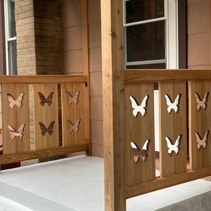 May include: A wooden porch railing with a decorative butterfly design. The railing is painted a light brown color and the butterflies are painted white.
