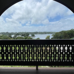 May include: A view from a balcony with a wooden railing featuring a decorative cutout design. The railing overlooks a body of water with trees and a cloudy sky in the background.