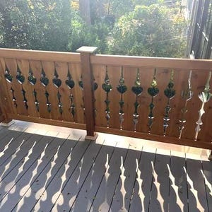 May include: A wooden deck railing with decorative cutouts in a repeating pattern. The railing is made of light brown wood and is installed on a deck with gray wooden planks. The sunlight casts shadows on the deck.